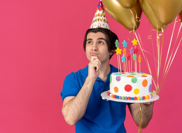 Confused handsome caucasian man wearing birthday cap puts hand on chin holds helium balloons and birthday cake 