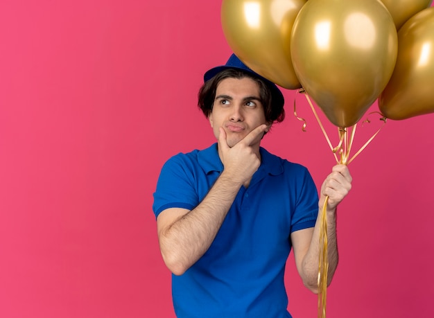 Confused handsome caucasian man wearing blue party hat holds helium balloons puts hand on chin 