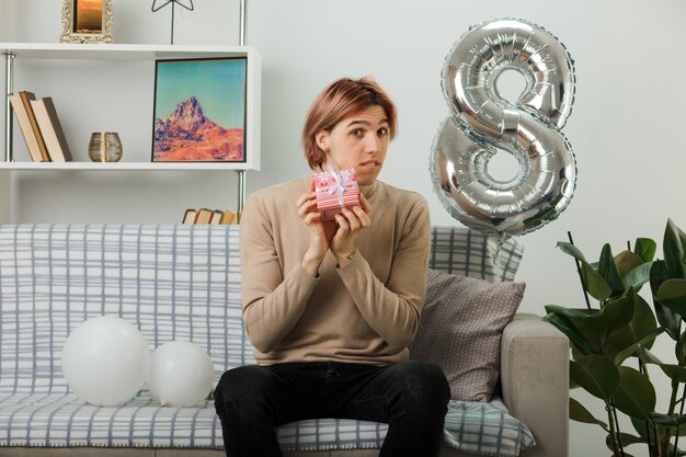 Confused handsome guy on happy women day holding present sitting on sofa in living room