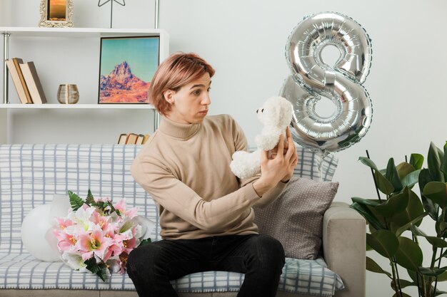 Бесплатное фото Смущенный красивый парень в счастливый женский день держит и смотрит на плюшевого мишку, сидящего на диване в гостиной