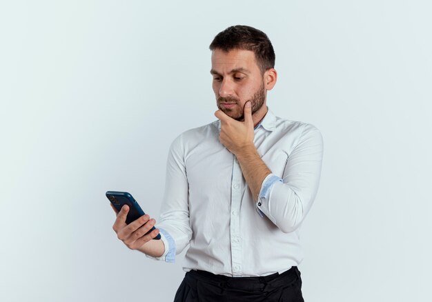 Confused handsome man puts hand on chin holding and looking at phone isolated on white wall