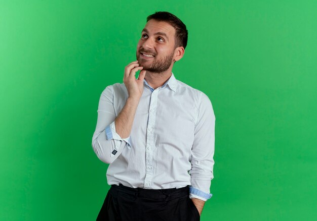 Confused handsome man puts hand on chin looking up isolated on green wall