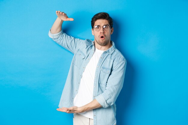 Confused handsome man showing big size, shaping large box and looking amazed, standing over blue background