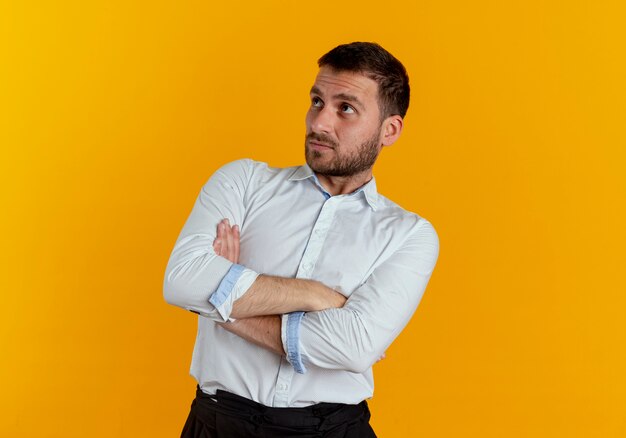 confused handsome man stands with crossed arms looking at side isolated on orange wall
