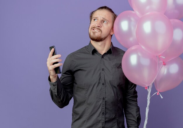 Confused handsome man stands with helium balloons holding phone looking up isolated on purple wall