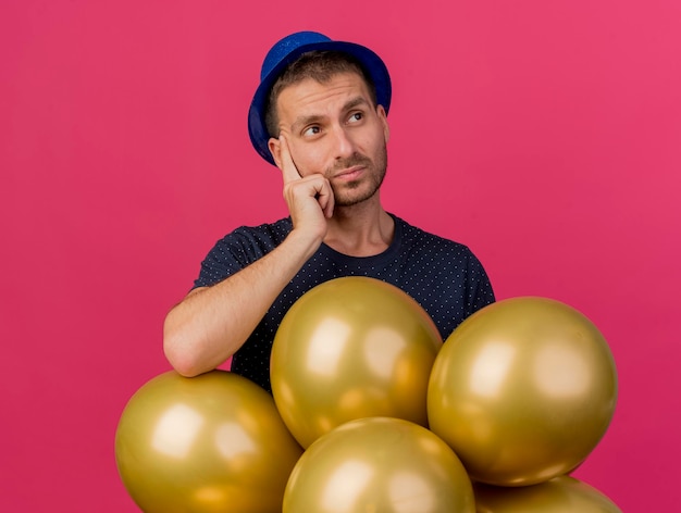 Uomo bello confuso che indossa cappello blu del partito tiene palloncini di elio guardando il lato isolato sulla parete rosa con lo spazio della copia