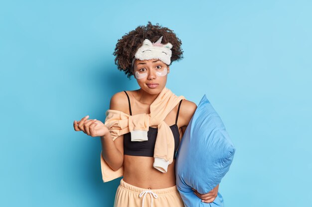 Confused hesitant young woman in pajama shrugs shoulders and feels uncertain about something holds pillow doesnt know plans for day stands indoor against blue wall