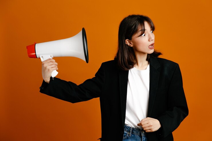 무료 Photo 오렌지 배경에 고립 된 검은 재킷을 입고 확성기를 들고 혼란스러운 젊은 아름다운 여성