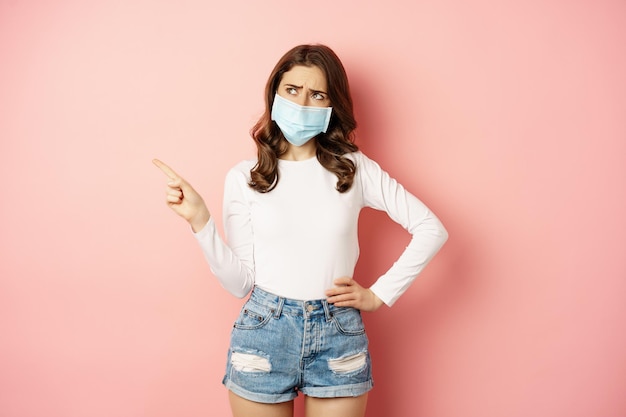 Free photo confused indecisive young woman in medical face mask pointing finger left and looking complicated ma...