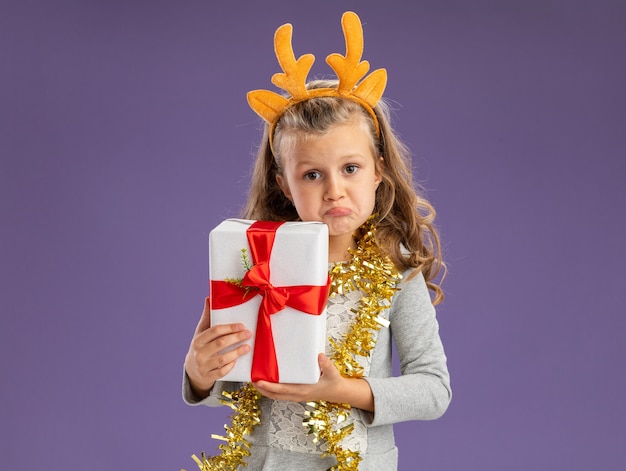 青い背景で隔離のギフトボックスを保持している首に花輪とクリスマスの髪のフープを身に着けている混乱した少女