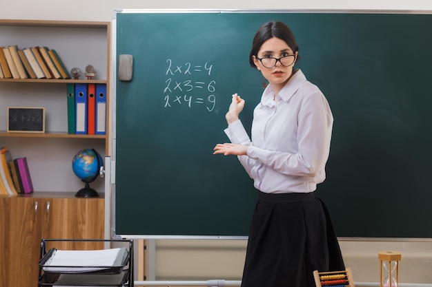 confused looking at camera young female teacher standing in front blackboard and write in classroom