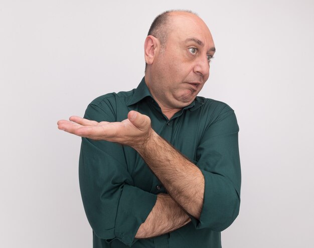 Confused looking at side middle-aged man wearing green t-shirt holding out hand at camera isolated on white wall