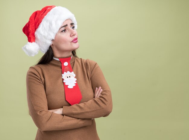 コピースペースでオリーブグリーンの背景に分離されたクリスマスの帽子とネクタイ交差点の手を身に着けている側の若い美しい少女を見て混乱