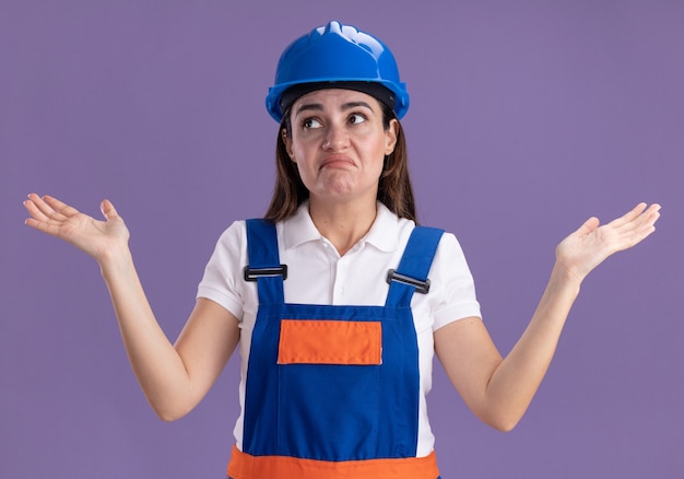 Foto gratuita confuso guardando lato giovane costruttore donna in uniforme diffondendo le mani isolate sulla parete viola