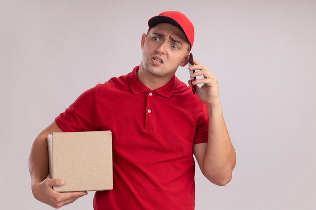 Confused looking at side young delivery man wearing uniform with cap holding box speaks on phone isolated on white wall with copy space