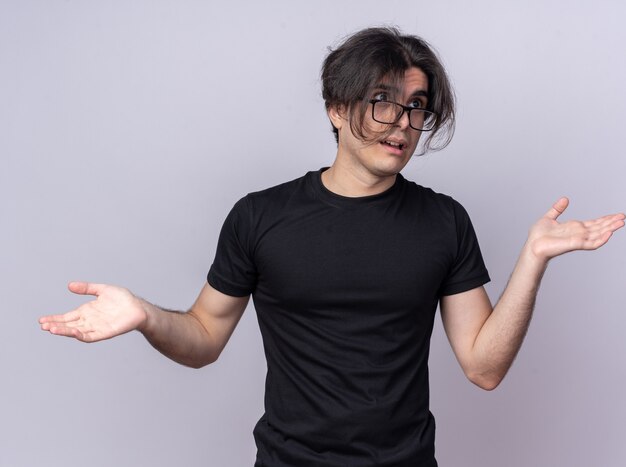 Confused looking at side young handsome guy wearing black t-shirt and glasses spreading hands isolated on white wall