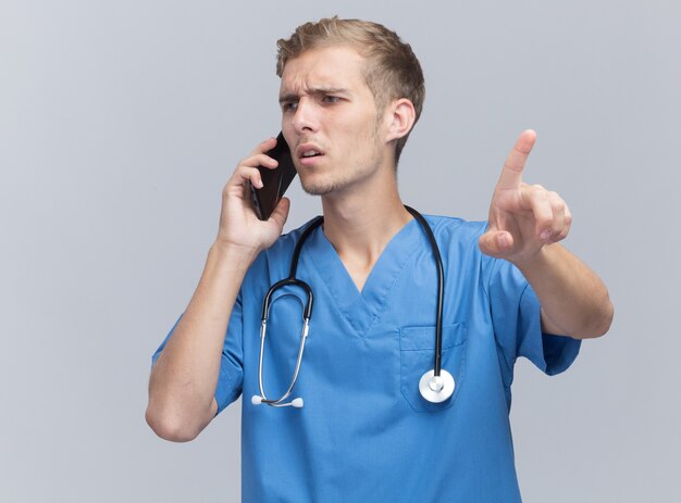 Free photo confused looking at side young male doctor wearing doctor uniform with stethoscope speaks on phone points at camera isolated on white wall