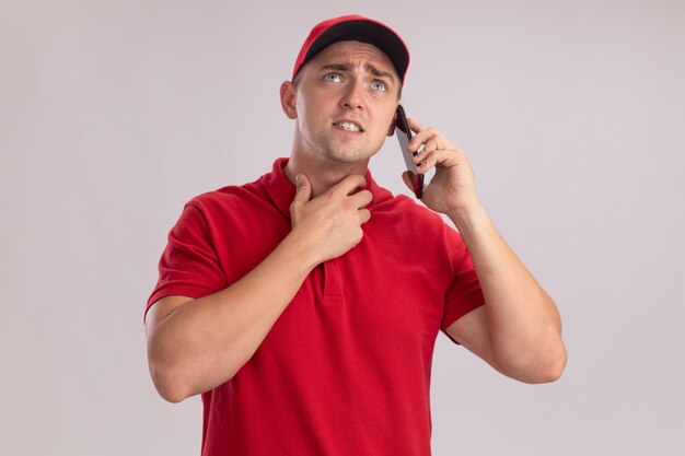 Confused looking up young delivery man wearing uniform with cap speaks on phone isolated on white wall