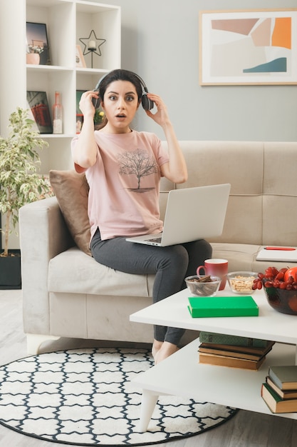 Confused looking young girl with laptop wearing headphones sitting on sofa behind coffee table in living room