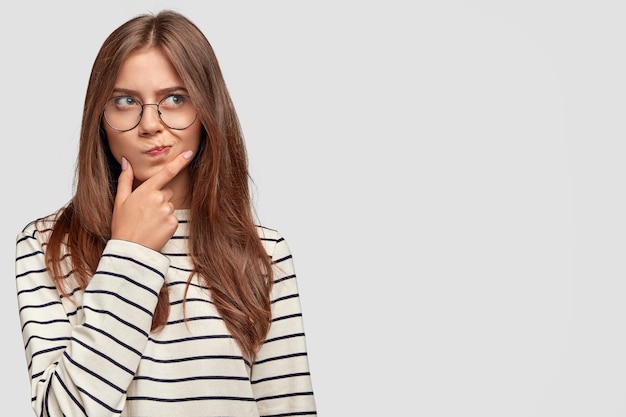 Confused lovely female teenager holds chin, looks thoughtfully aside, has dark hair, wears striped sweater, isolated over white wall