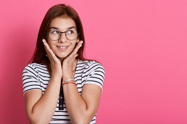 Confused lovely female teenager looks thoughtfully aside, has dark hair, wears striped t shirt, posing isolated over pink wall, thoughtful woman indoor.