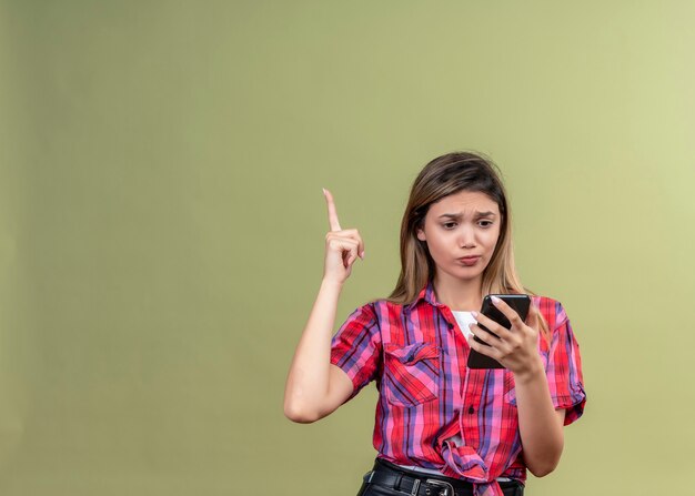 A confused lovely young woman in a checked shirt looking at mobile phone while raising hand on a green wall