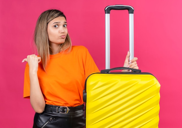 ピンクの壁に黄色のスーツケースを保持しているオレンジ色のTシャツの混乱した素敵な若い女性