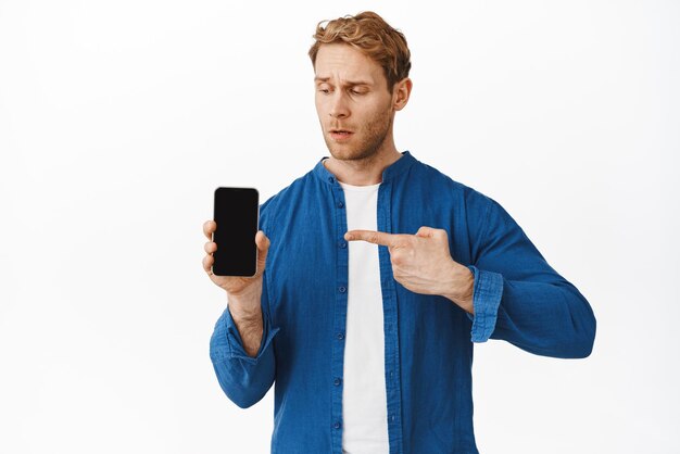 Confused man asking question about smartphone app pointing at mobile phone screen and look interested intrigued by new feature shopping offer standing over white background