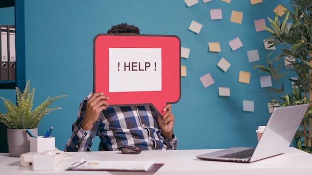 Confused man holding speech bubble to ask for help, working on laptop for executive business. Person using card board banner with text message on camera, showing communication symbol.