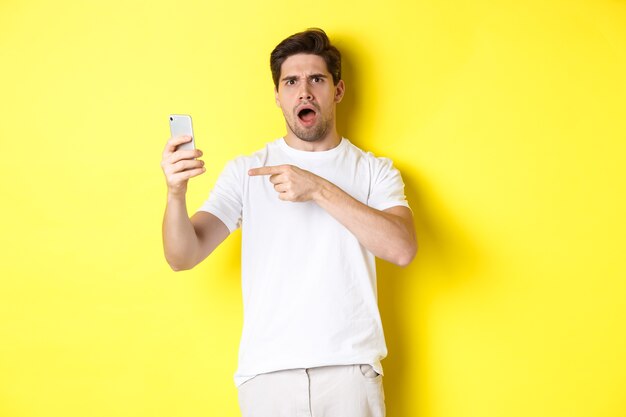 Confused man pointing finger at mobile phone screen, cant understand something, standing over yellow background. Copy space