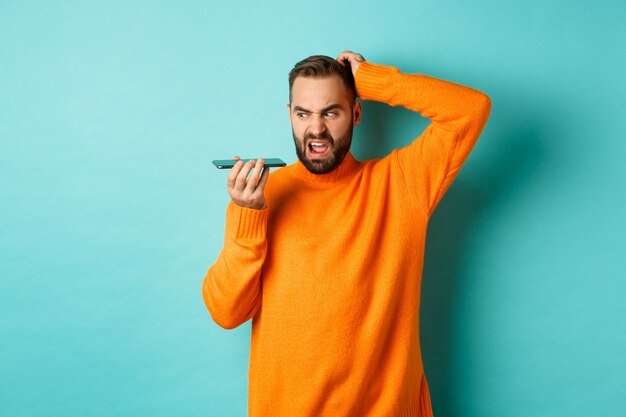 Confused man scratching head while talking on speakerphone, record voice message with indecisive face, standing in orange sweater over light turquoise wall.