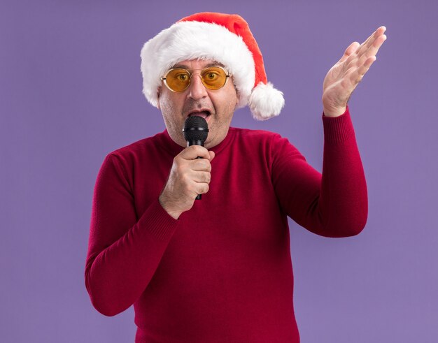 紫色の背景の上に立って腕を上げてマイクに向かって話している黄色いメガネでクリスマスサンタの帽子をかぶって混乱した中年男性