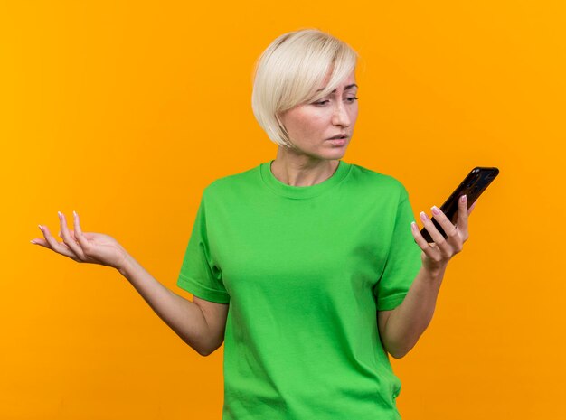 Confused middle-aged blonde slavic woman holding and looking at mobile phone isolated on yellow wall
