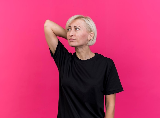 Confused middle-aged blonde slavic woman keeping hand behind head looking at side isolated on pink wall with copy space