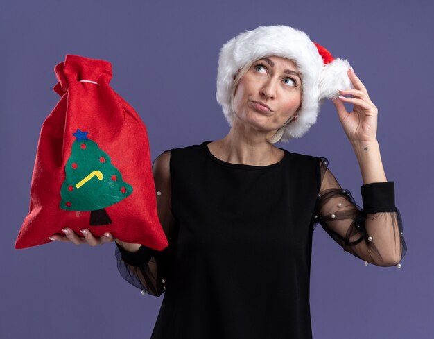 Confused middle-aged blonde woman wearing christmas hat holding christmas sack touching hat looking up isolated on purple background