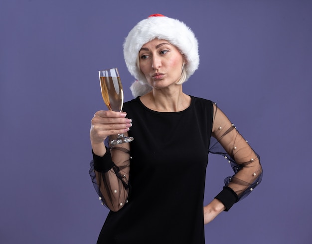 Confused middle-aged blonde woman wearing christmas hat holding and looking at glass of champagne keeping hand on waist isolated on purple background with copy space