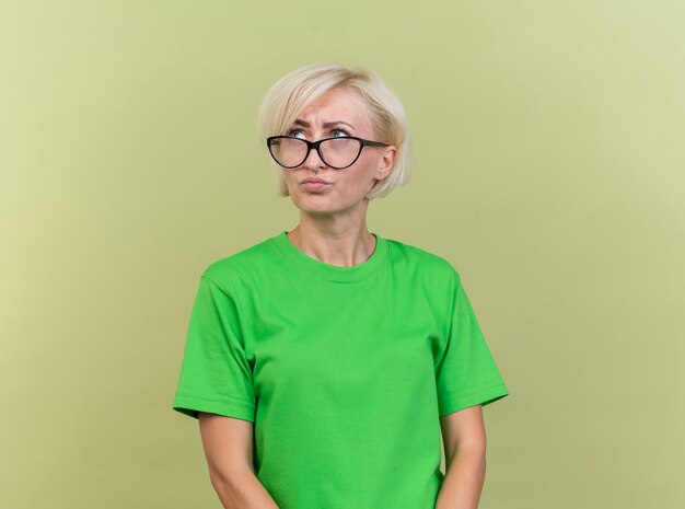 Смущенная блондинка средних лет в очках смотрит в сторону, изолированную на оливково-зеленой стене