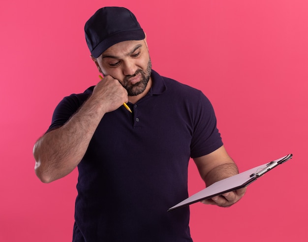 Confused middle-aged delivery man in uniform and cap holding and looking at clipboard putting fist on cheek isolated on pink wall