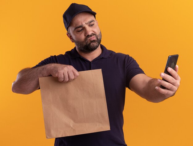 Confused middle-aged delivery man in uniform and cap holding paper food package and looking at phone in his hand isolated on yellow wall