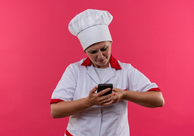 격리 된 분홍색 벽에 그녀의 손에 전화를보고 요리사 유니폼에 혼란 중년 여성 요리사