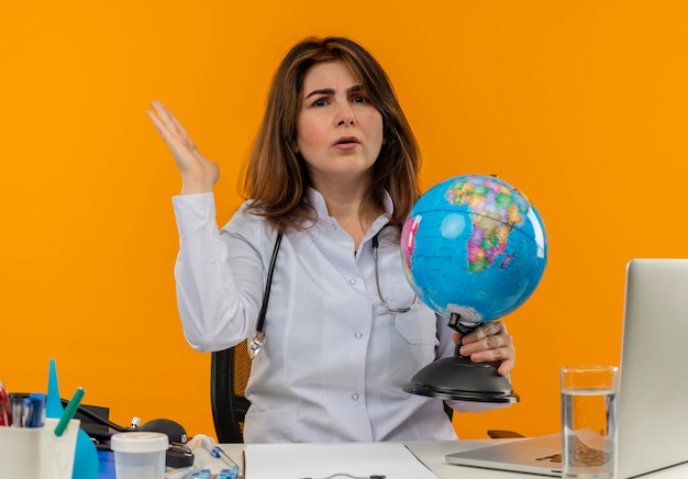 Confused middle-aged female doctor wearing medical robe with stethoscope sitting at desk work on laptop with medical tools holding globe and spread hand on isolated orange wall with copy space