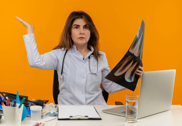 Confused middle-aged female doctor wearing medical robe with stethoscope sitting at desk work on laptop with medical tools holding x-ray and raising hand on isolated orange wall