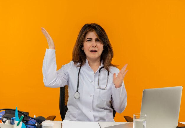 Confused middle-aged female doctor wearing medical robe with stethoscope sitting at desk work on laptop with medical tools raising hands on isolated orange wall with copy space