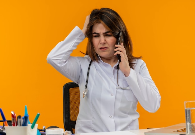 Confused middle-aged female doctor wearing medical robe with stethoscope sitting at desk work on laptop with medical tools speaks on phone and putting hand on head on orange wall with copy space