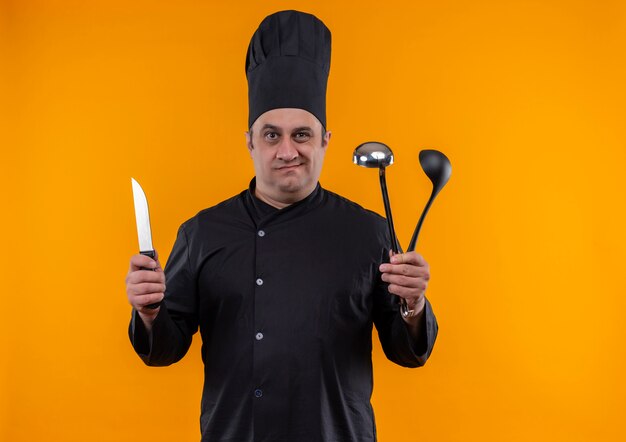 Confused middle-aged male cook in chef uniform holding ladle and knife on yellow wall with copy space