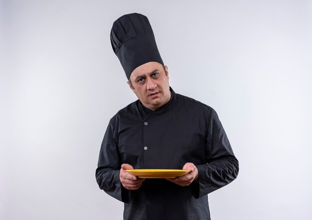 Confused middle-aged male cook in chef uniform holding plate on isolated white wall