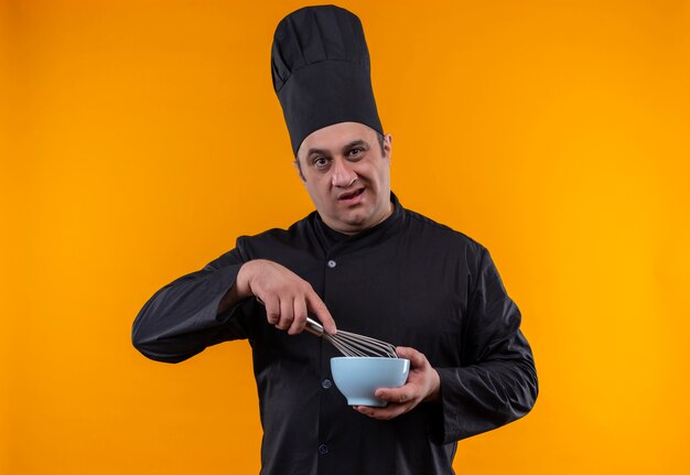 Confused middle-aged male cook in chef uniform holding whisk and bowl on yellow wall