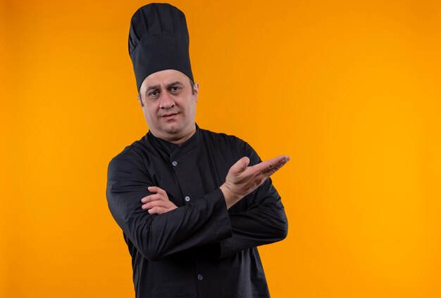 Confused middle-aged male cook in chef uniform points hand to side on yellow wall with copy space