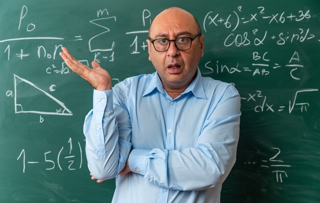 Confused middle-aged male teacher wearing glasses standing in front blackboard spreading hand