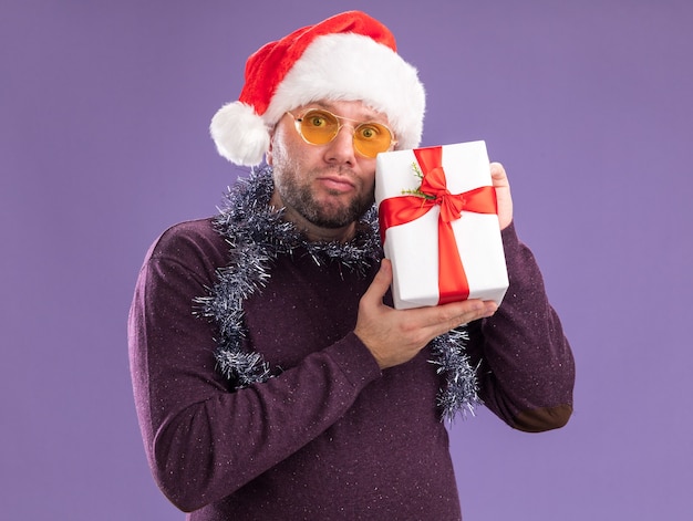 보라색 벽에 고립 된 선물 패키지를 들고 안경 목에 산타 모자와 반짝이 갈 랜드를 입고 혼란 중년 남자